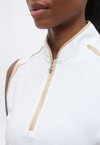 Κοντινό πλάνο ενός ατόμου που φοράει λευκό αμάνικο τοπ με μπεζ φάσα και ημικοντό φερμουάρ στον λαιμό.