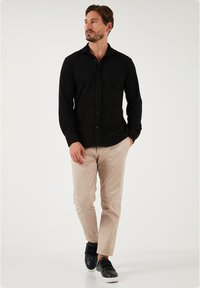 Camisa negra de manga larga con botones, pantalones beige de corte ajustado y zapatillas negras con tiras de velcro. Modelo de pie contra un fondo blanco.