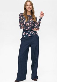 Donna sorridente, con indosso una blusa a maniche lunghe trasparente con motivi floreali e pantaloni con gamba larga di un blu scuro, in piedi di fronte a uno sfondo bianco.