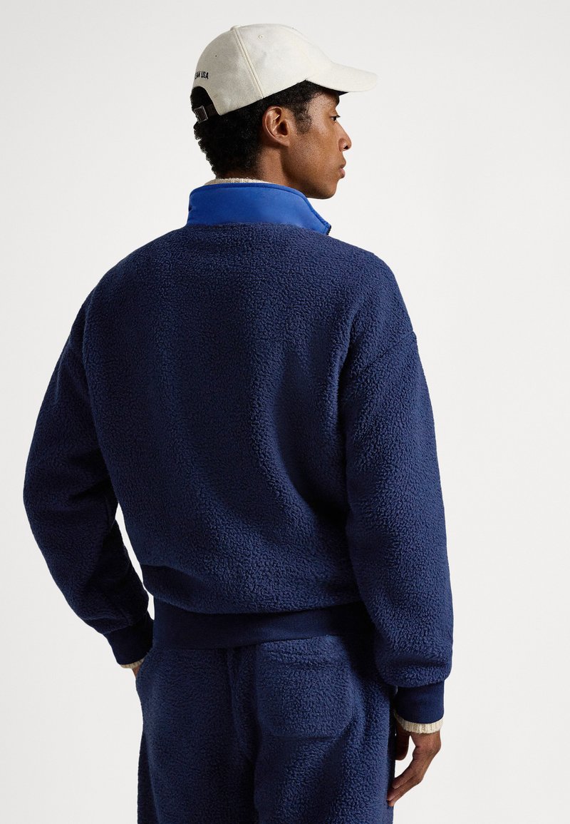 Pull polaire bleu marine avec col montant, surface texturée, poignets et ceinture côtelés. Associé à un pantalon assorti bleu marine et une casquette blanche.