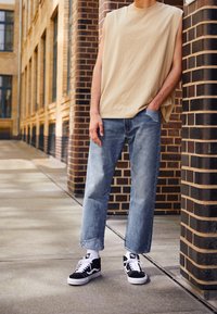 Beige ärmlös skjorta, ljusblå raka jeans och svarta sneakers med vita sulor; avslappnad streetwear-estetik.