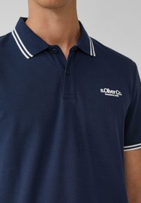 Marineblaues Poloshirt mit weißem gestreiftem Kragen und s.Oliver-Logo. Texturiertes Gewebe mit drei Knöpfen an der Knopfleiste. Klassische Passform.