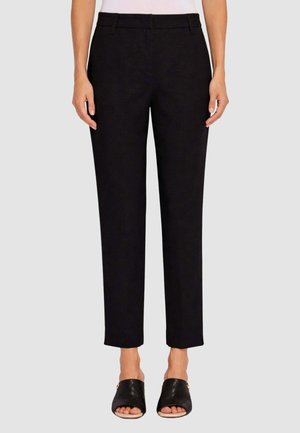 Donna che indossa pantaloni neri su misura fino alla caviglia e scarpe nere aperte senza lacci, in piedi contro uno sfondo semplice.