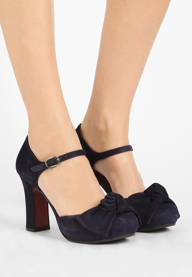 Chaussures à talons hauts en daim bleu marine avec brides à la cheville et nœuds décoratifs à l'avant, portées pieds nus.