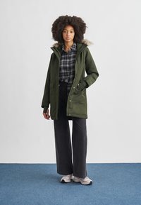 Olive green parka with a fur-lined hood, zip and snap closure, side pockets, and a relaxed fit, paired with black wide-leg pants and sneakers.