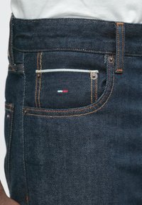 Σκούρα μπλε τζιν από τζιν με πορτοκαλί ραφές, που διαθέτουν μια μικρή μπροστινή τσέπη με κόκκινη, λευκή και μπλε λεπτομέρεια λογότυπου �και μεταλλικά καρφιά.