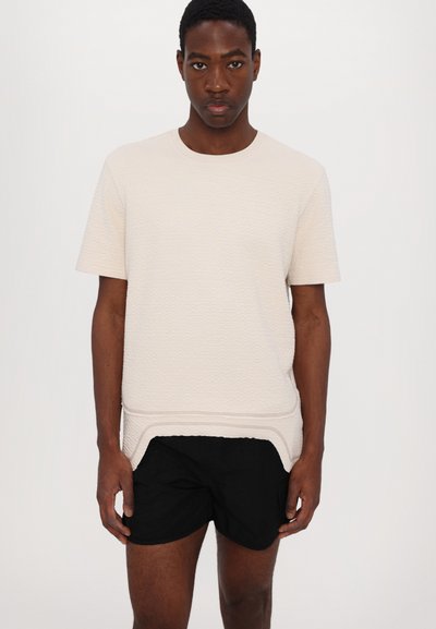Korte mouwen, lichtbeige textuurshirt met een gebogen zoom over zwarte shorts. Glad materiaal met subtiele lijnen en een ontspannen pasvorm.