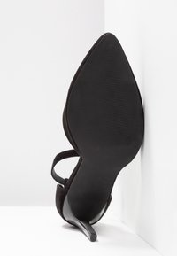 Chaussure à talon haut noire à bout pointu avec talon stiletto fin, positionnée debout contre un mur d'angle blanc.