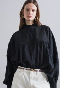 Zwarte oversized blouse van textuurstof, met plooien en geborduurde accenten, gecombineerd met witte broeken en een riem met gouden accenten.