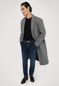 Grå ullkappa över en svart tröja, kombinerat med blå denimjeans och mörkbruna skor. Funktioner inkluderar fickor och en elegant silhuett.