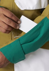 Kolorowa kurtka z zielonym rękawem, wyposażona w kieszonkę na kartę. Teksturowana tkanina z mieszanką żółtych i białych elementów.