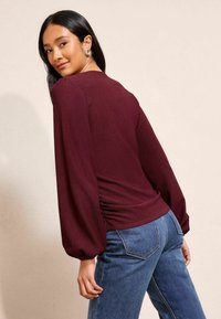 Burgunderfarbene Langarmbluse aus strukturiertem Stoff mit Raffung an der Seite, kombiniert mit mittelblauen Jeans aus Denim, die klassische Gesäßtaschen haben.