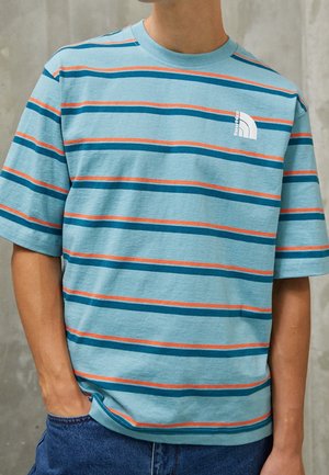 Mężczyzna ubrany w niebieski T-shirt z krótkim rękawem w turkusowe i pomarańczowe poziome paski oraz małym białym logo na piersi.