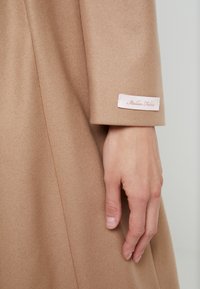 Main reposant sur le côté, portant un manteau en laine couleur chameau avec une petite étiquette indiquant "Italian Fabric" sur le poignet de la manche.
