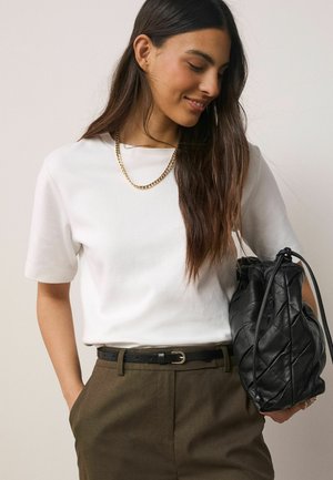 Pikkade tumedate juustega naine, kes kannab valget lühikeste varrukatega särki, kuldse kettkaelakeed, pruune pükse musta vööga ja hoiab musta õmmeldud kotti.