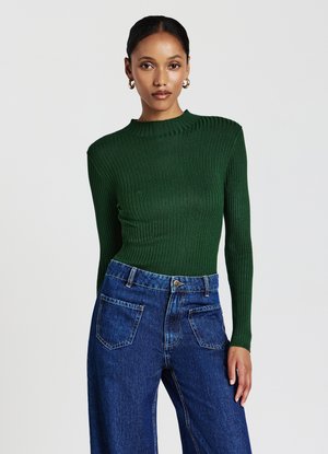Maglietta lunga a maniche con costine verdi e colletto alto, abbinata a jeans in denim blu a vita alta con grandi tasche posteriori.