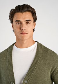 Groene cardigan met een geribbelde kraag en knoopsluiting, gedragen over een eenvoudig wit T-shirt, met een gebreid textuurpatroon.