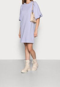 Femme portant une robe lilas à manches bouffantes, des bottines beiges à lacets et tenant un petit sac à bandoulière beige, se tenant sur un sol gris.