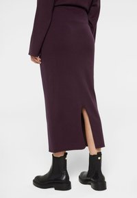 Violette Strick-Midirock mit einem Rückenschlitz, detaillierter Nahtführung und glatter Textur, kombiniert mit schwarzen Ankle Boots mit goldenen Akzenten.