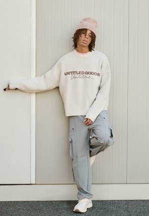 Crème trui met de tekst "UNTITLED GOODS", lichtgrijze cargopants en witte sneakers. Roze beanie voegt een zachte accent toe. Glad textuur.