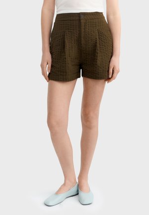 Kobieta nosząca ciemnozielone, wysokiej talii szorty z teksturą, biały top i jasnoniebieskie płaskie buty, stojąca z rękami swobodnie opuszczonymi wzdłuż ciała.