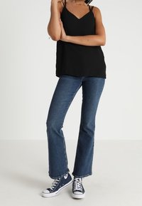 Vrouw in een zwarte mouwloze top, blauwe flared jeans en marineblauwe high-top sneakers, staand met gekruiste benen.