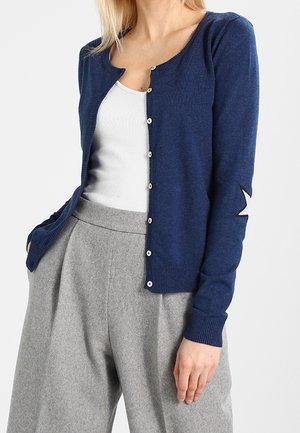 Žena v tmavě modrém kardiganu na knoflíky s bílou hvězdou na rukávu, bílém tílku a šedých širokých kalhotách s vysokým pasem.