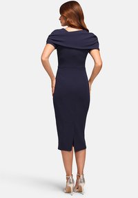 Navyblaues, tailliertes Kleid mit drapiertem Ausschnitt, knielanger Länge und einem Schlitz hinten in der Mitte. Kombiniert mit metallic-farbenen, rutschfesten High Heels.