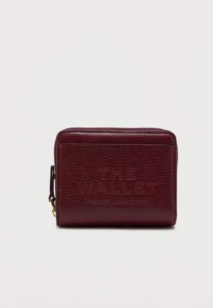 Portofel din piele bordeaux, cu text embossat "THE WALLET" și "MARC JACOBS." Dispune de un compartiment cu fermoar și o suprafață texturată. Design compact.