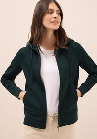 Felpa con zip di colore verde scuro, con cappuccio regolabile, polsini a costine e due tasche frontali, indossata sopra una t-shirt bianca e pantaloni beige.
