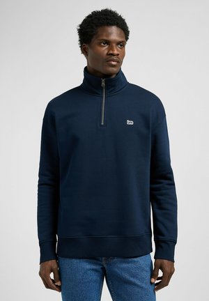 Navyblauwe ritsvaste sweatshirt met hoge kraag, ribgebreide manchetten en zoom. Voorzien van een klein wit logo op de borst. Gemaakt van zacht materiaal.