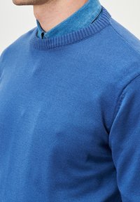 Maglione blu lavorato a maglia con scollo a costine, caratterizzato da una texture liscia e un design aderente, indossato sopra una camicia color azzurro chiaro con colletto.