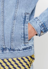 Veste en denim bleu clair avec une main dans la poche latérale, bouton de poignet visible, sur un tissu à motif zigzag jaune et bleu.
