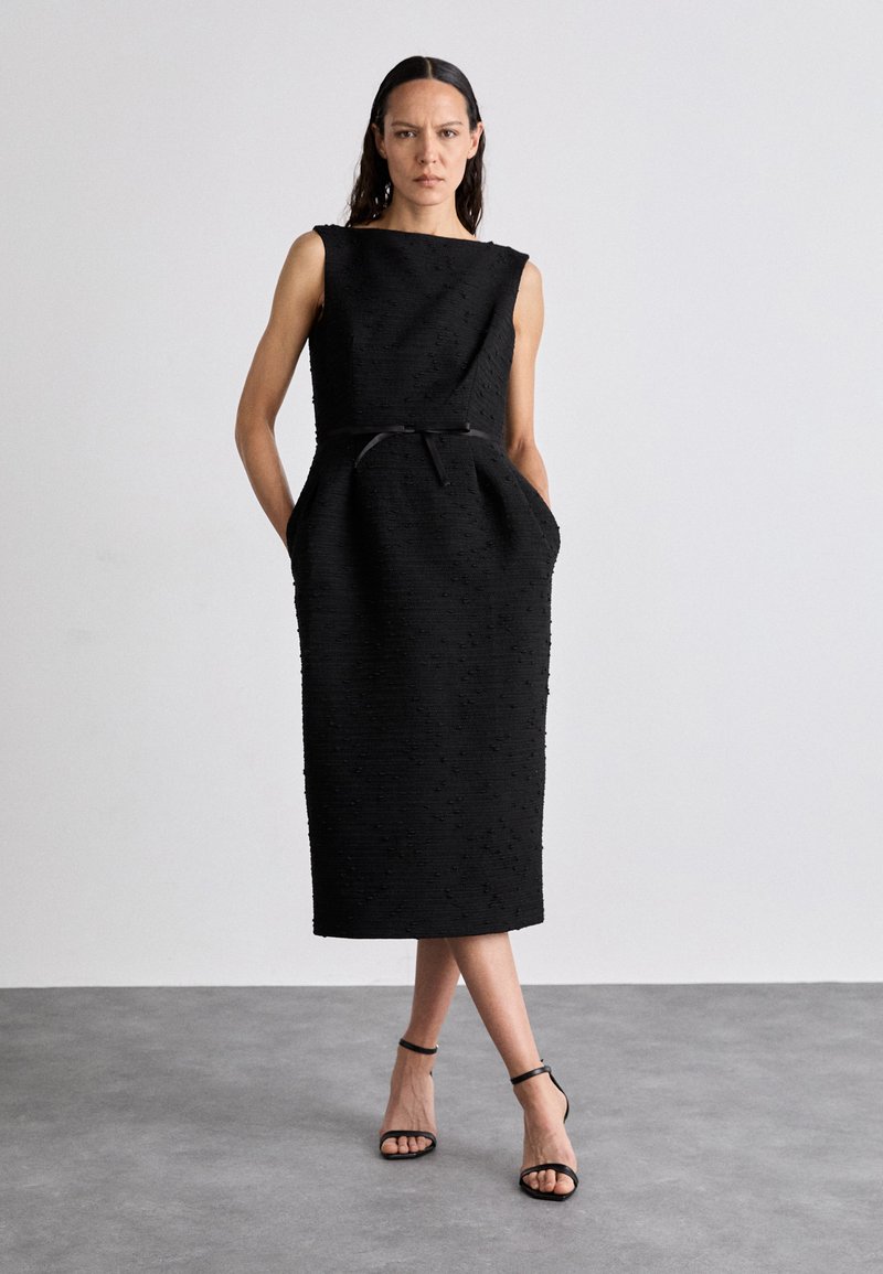 Svart ärmlös klänning med texturerad tyg, knälång, med en strukturerad silhuett, sidofickor och ett smalt bälte i midjan.