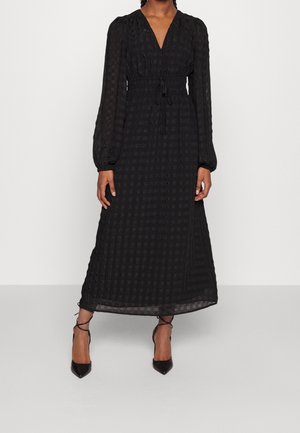 Woman wearing a long black checkered dress with long sleeves, a V-neck, and a waist tie, paired with black ankle-wrap heels.