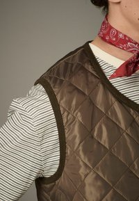 Brown quilted vest with olive trim, worn over a white and black striped shirt. Red bandana tied around the neck.