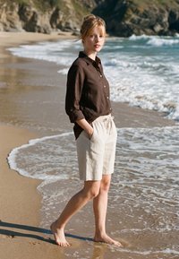 Jeune femme en chemise sombre et short clair, debout pieds nus sur du sable humide sur une plage avec des vagues et des falaises rocheuses en arrière-plan.