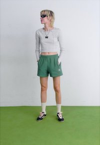 Λαμπερό γκρι μπλουζάκι με μακριά μανίκια και κοντή γραμμή, πράσινες βερμούδες με λευκό λογότυπο, λευκές κάλτσες μέχρι το γόνατο, μαύρα αθλητικά παπούτσια με ροζ κορδόνια, κοντά ξανθά μαλλιά.