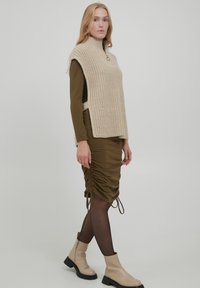 Beige gestricktes ärmelloses Oberteil mit hohem Kragen über einem taillierten olivgrünen Kleid mit Raffung, kombiniert mit hellbeigen Stiefeletten.