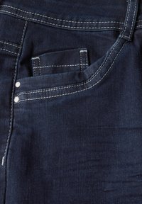 Dunkelblaue Denim-Jeans mit einer Vordertasche, weißen Nähten und metallischen Nieten für zusätzliche Details. Weiche Textur mit glatter Oberfläche.