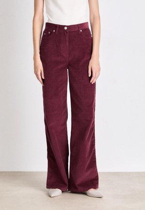 Woman wearing high-waist wide-leg burgundy corduroy pants with front pockets and a white top standing on a light floor.