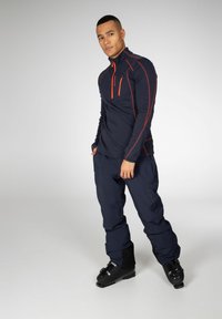 Maglia termica a maniche lunghe color navy con accenti arancioni; colletto alto; tasca con zip. Abbinata a pantaloni scuri e stivali da sci neri.