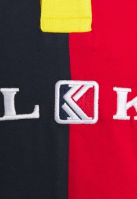 Puuvillapolo-shirt, jossa on mustia, punaisia ja keltaisia osia; sisältää brodeeratun logon brittiläisten lippuvärien sävyissä. Sileä rakenne.