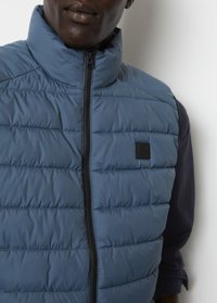Blaues, gepolstertes Westchen mit hohem Kragen, frontalem Reißverschluss und einem kleinen Markenlogo. Die Textur ist glatt und mit horizontalen Steppnähten verziert.