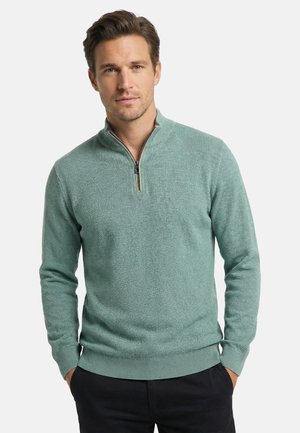 Uomo che indossa un maglione verde chiaro testurizzato con zip al quarto, mani nelle tasche, su uno sfondo chiaro e uniforme.