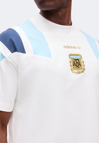 Άνδρας που φοράει λευκό μπλουζάκι Adidas με γαλάζιες και σκούρες μπλε ρίγες στους ώμους και χρυσό έμβλημα της Ποδοσφαιρικής Ομοσπονδίας Αργεντινής.