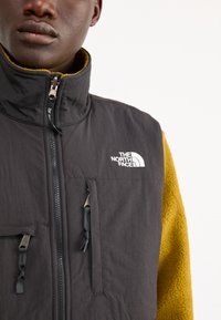 Gilet isolato nero con colletto alto, chiusura zip e logo. Presenta due tasche con zip e un rivestimento in pile giallo testurizzato sotto.