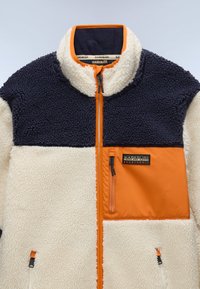 Cream and navy fleece jacket with orange accents, zippered pockets, high collar, and logo patch. Features a textured, plush fabric.