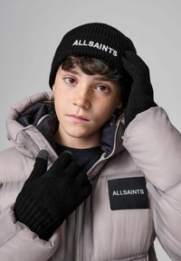 Černý pletený klobouk s logem "ALLSAINTS", spárovaný s černými pletenými rukavicemi; model nosící světle šedou péřovou bundu s logem na náplasti.