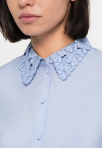 Lyseblå skjorte med brodert blomsterkrage. Kragen har intrikate mønstre, og stoffet har en glatt tekstur med knappelukking.